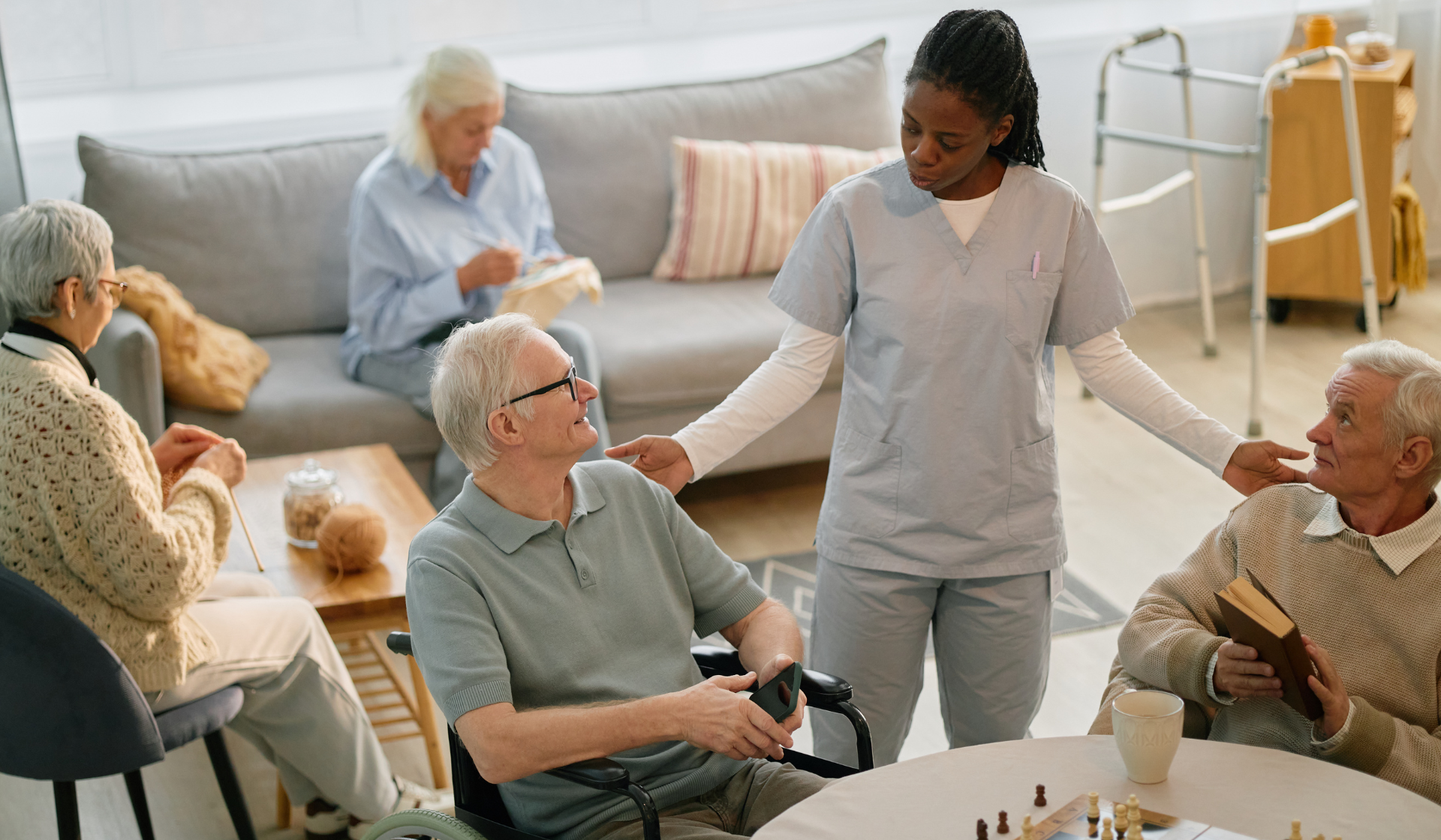 senior aid talking to two senior men at an assisted living facility, with two seniors behind them knitting